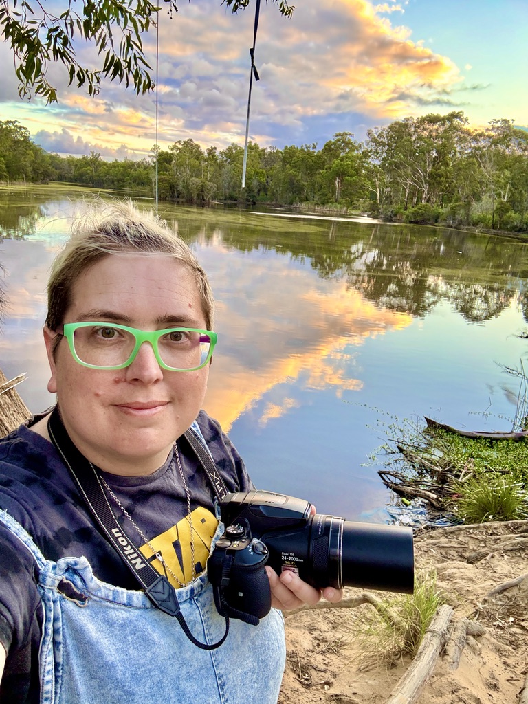 Jess holding a Nikon P900 camera by a dam at sunset, reflections on the water